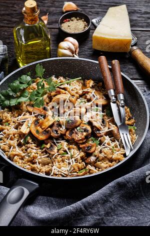 risotto classico cremoso ai funghi cosparso di parmigiano grattugiato in padella e ingredienti su un tavolo di legno scuro, vista verticale, cucina italiana Foto Stock