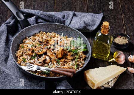 classico risotto cremoso ai funghi cosparso di parmigiano grattugiato in una padella e ingredienti su un tavolo di legno scuro, cucina italiana Foto Stock