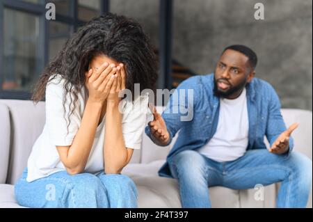 Concetto di abuso domestico. Donna multirazziale depressa si siede sul divano e piange, un uomo nero irritato e arrabbiato sta gridando sullo sfondo. Una coppia sta discutendo a casa Foto Stock