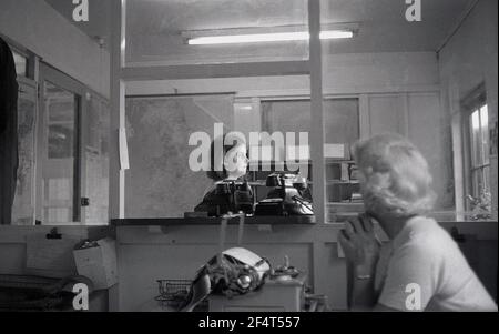 1964, storico, daydreaming.... all'interno dei locali di un'azienda industriale, una donna matura in un ufficio parted seduto ad una macchina da scrivere che guarda attraverso la finestra partizionata ad un'altra, donna più giovane nell'ufficio accanto in piedi da due grandi vecchi telefoni bakerlite dell'era che guarda fuori, Inghilterra, Regno Unito. Foto Stock