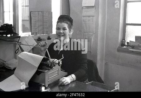 1964, storico, all'interno di un ufficio di una società commerciale che vende rotavators e coltivatori, una lavoratrice chierica femminile seduta ad una scrivania di legno ad una macchina da scrivere, con una grande stampa fuori in, Inghilterra, Regno Unito. Foto Stock