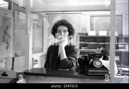 1964, storico, all'interno di un ufficio divisionato con pannelli di vetro presso una società di ingegneria leggera, con grandi mappe sulle pareti, una giovane donna membro del personale, in piedi al banco salari o boccata accanto a un grande telefono bakerlite, Inghilterra, Regno Unito. Foto Stock