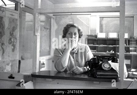 1964, storico, all'interno di un ufficio partizionata in vetro pannellato presso una società di ingegneria leggera, una donna matura membro del personale, in piedi al banco di stipendi o portello accanto a un grande telefono bakerlite, Inghilterra, Regno Unito. Foto Stock