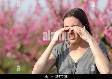 Donna stressata graffiando gli occhi pruriginosi in primavera in un rosa campo di fiori Foto Stock