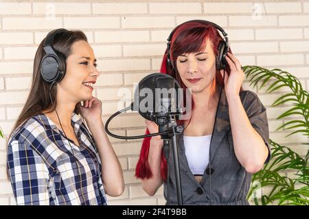 Felice giovane femmina in cuffia sorridente e ascoltando la ragazza canto con occhi chiusi mentre si registra musica contro mattoni parete in studio Foto Stock