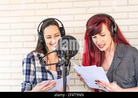 Felice giovane attrici voce in cuffie sorridenti e lettura script vicino al microfono durante la creazione di colonna sonora per film contro pareti in mattoni Foto Stock