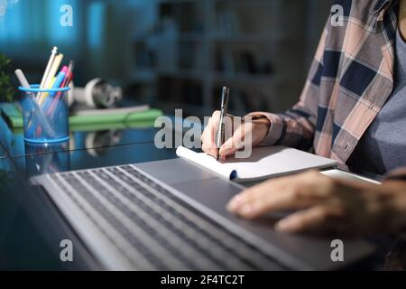 Primo piano di una mano dello studente che prende appunti controllando il laptop di notte a casa Foto Stock