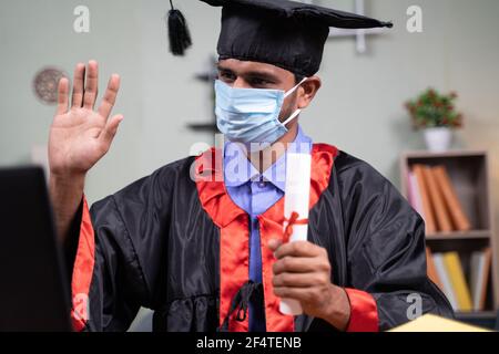 Studente con maschera medica che partecipa a una cerimonia virtuale di laurea video da casa su laptop: concetto di assistenza sanitaria, sicurezza e novità normale Foto Stock