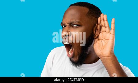 Divertente Black Guy ascolto tenere la mano vicino all'orecchio, sfondo blu Foto Stock