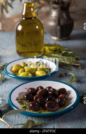 Olive naturali nere e verdi con rosmarino, basilico e foglie di olivo su sfondo di legno blu. Foto Stock
