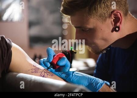 Il tatuaggio principale disegna la vernice nera sul tatuaggio del cliente. Artista del tatuaggio che tiene una macchina per tatuaggio in guanti sterili blu e lavora su Foto Stock