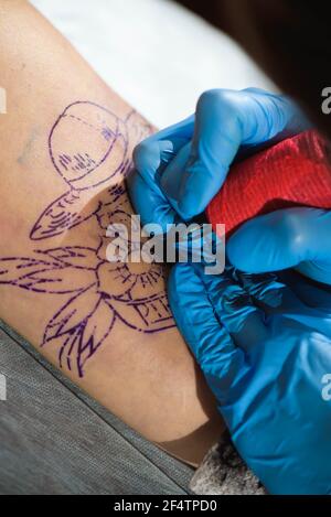 Il tatuaggio principale disegna la vernice nera sul tatuaggio del cliente. Artista del tatuaggio che tiene una macchina per tatuaggio in guanti sterili blu e lavora su Foto Stock