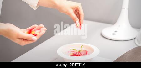 Le mani femminili dell'estetista preparano il bagno di manicure con il rosso e il rosa petali di rose sul tavolo in spa Foto Stock