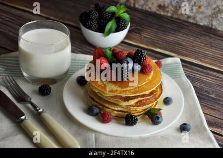 Frittelle soffici fatte in casa, posate, latte e una ciotola di frutti di bosco sullo sfondo. Foto Stock