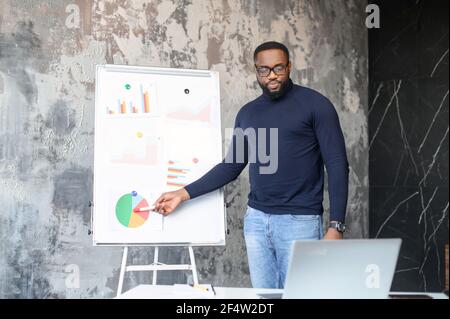 Un impiegato di sesso maschile afro-americano intelligente e contemporaneo che tiene una videoconferenza online, un ragazzo nero sicuro punta ai grafici sulla lavagna, spiegando smth al pubblico online sul laptop Foto Stock