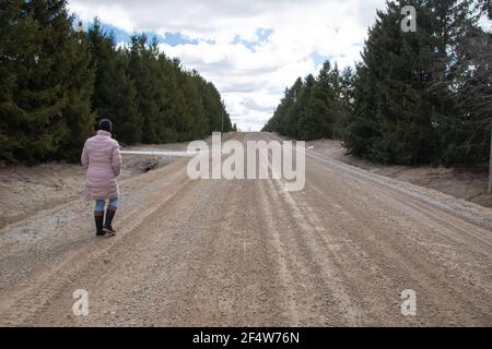 Una donna che indossa un parka di puff rosa e una punta nera e jeans cammina lungo una vecchia strada sterrata nella campagna fiancheggiata da alberi di abete rosso conifero. Foto Stock