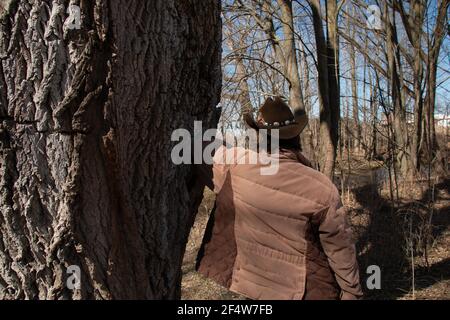 Una donna con capelli corti marroni che indossa un cappello cattleman cowboy con una piuma e cintura decorativa attaccata. Appoggiandosi ad un albero Foto Stock