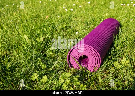 Tappeto fitness arrotolato su erba verde con luce solare, primo piano. Yoga all'aperto Foto Stock