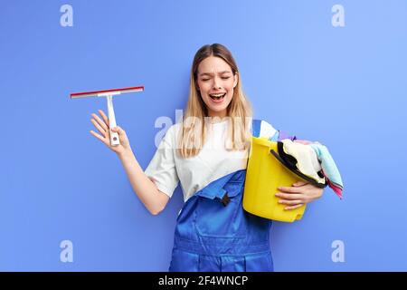 piangendo femmina con i rifornimenti di pulizia in piedi gridando da sporcizia in camera deve pulire, isolato su sfondo viola. ritratto di cameriera in tute Foto Stock