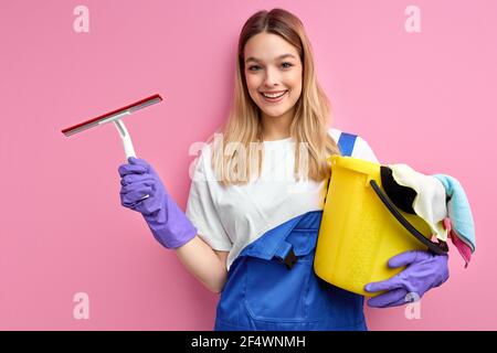 Donna sorridente casalinga in guanti che tengono le cose più pulite isolato su sfondo rosa studio. Ritratto di bella donna pronta a pulire la camera sporca, enj Foto Stock