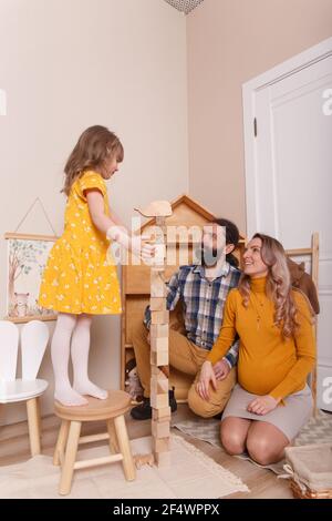 La famiglia sta aspettando una nuova aggiunta. Una donna incinta, suo marito e sua figlia giocano insieme nella stanza dei bambini. Costruiscono una torre di legno bl Foto Stock