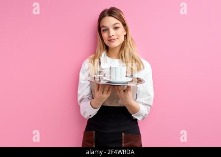 Cameriera che offre una tazza di caffè isolato su sfondo rosa, sorridente signora in uniforme dare tazza di bevanda al cliente, indossando maschera medica. Verticale Foto Stock