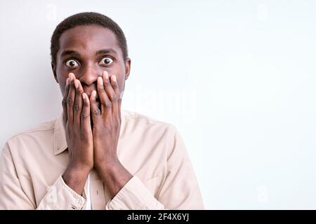 l'uomo africano scioccato si trova nella paura, spaventato da qualcosa, chiudendo la bocca e guardando la macchina fotografica, isolato su sfondo bianco Foto Stock