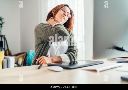 Una donna di mezza età con dolore al collo mentre lavora come graphic designer utilizzando un computer moderno nel suo soggiorno. Lavoro a distanza o freelance su w Foto Stock