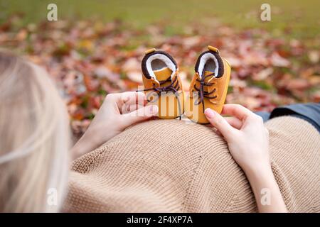 Donna incinta con le scarpe cute del bambino nel parco autunnale, closeup. Futura madre che tiene le scarpe del bambino sulla sua pancia incinta. Concetto di genitori-essere. Foto Stock