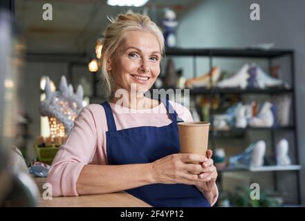 Ritratto di felice donna matura in grembiule blu sorridente a macchina fotografica, bere caffè o tè da tazza usa e getta nel suo studio di ceramica Foto Stock