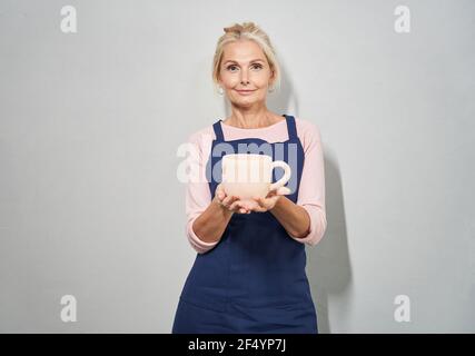 Ritratto di bella donna matura in grembiule blu sorridente a macchina fotografica, mostrando la tazza di ceramica di argilla fatta a mano da lei in ceramica, che posa su bianco Foto Stock