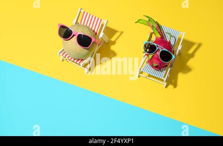 Vista dall'alto con frutta a baldacchini e draghi seduti su sedie da spiaggia in miniatura con occhiali da sole. Concetto estivo con frutta esotica per prendere il sole al Foto Stock