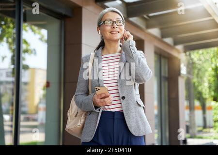 Smiley donna dai capelli grigi che indossa cuffie wireless durante la sua passeggiata Foto Stock