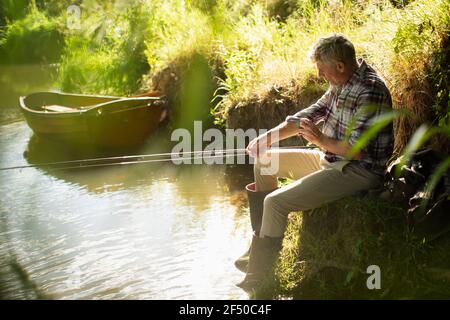 L'uomo pesca a mosca rotolando le maniche sulla soleggiata riva del fiume Foto Stock