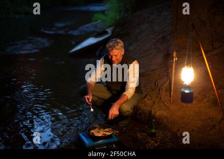 Pescatore maschio che cucinano pesce fresco catturato su stufa da campeggio a. notte Foto Stock