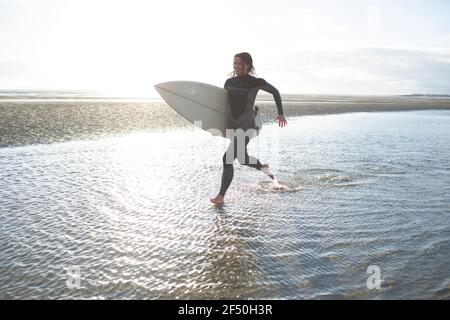 Giovane surfista femminile che corre con la tavola da surf nella soleggiata spiaggia dell'oceano surf Foto Stock