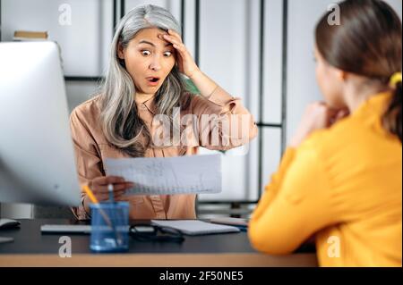 Scioccata indignata donna asiatica matura con capelli grigi, leader aziendale, top manager, capo, in un ufficio moderno, in abiti eleganti, guarda i documenti del dipendente con sorpresa, in orrore Foto Stock