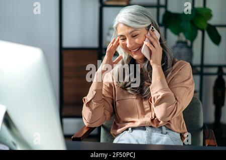 Una donna d'affari asiatica con capelli grigi, un ceo, che lavora alla scrivania, indossa abiti eleganti, parla con i colleghi o gli amici su smartphone, sorridendo amichevole Foto Stock