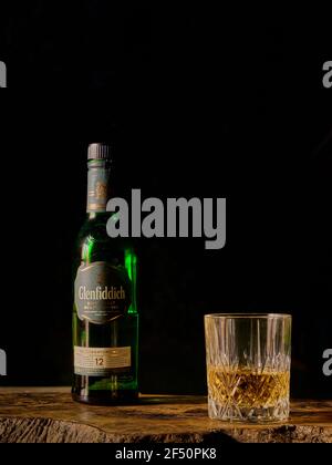 Glenfiddich Scotch Whisky bottiglia e vetro mezzo riempito su un asse di legno di oliva su uno sfondo nero scuro Foto Stock