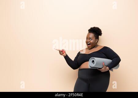 donna afro-americana soprappeso stupita in abbigliamento sportivo con tappetino fitness e puntando con il dito isolato sul beige Foto Stock