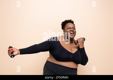 allegra donna afroamericana plus taglia in abbigliamento sportivo con manubri isolato su beige Foto Stock