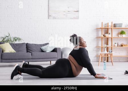 vista laterale della donna afroamericana plus size in abbigliamento sportivo esercitarsi sul tappetino di idoneità in soggiorno Foto Stock
