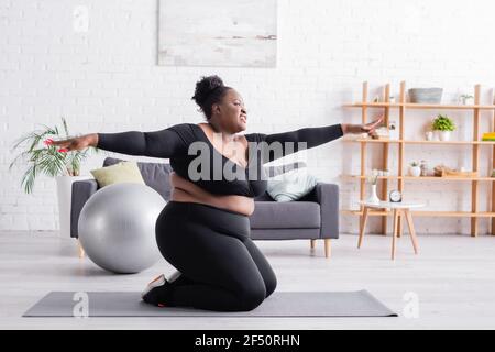 felice african american plus taglia donna in abbigliamento sportivo con outtensed mani che si esercitano in soggiorno Foto Stock