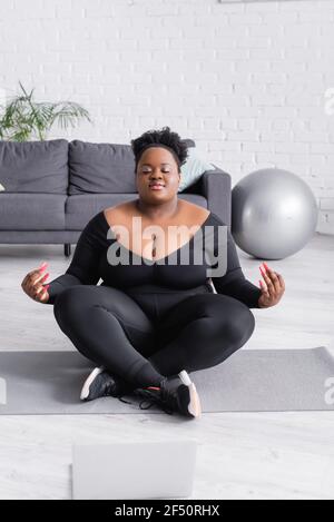 donna afroamericana più taglia in abbigliamento sportivo seduto in yoga mettiti in posa vicino al computer portatile nel soggiorno Foto Stock