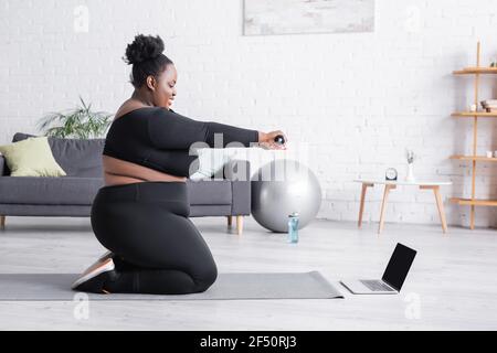 vista laterale di african american più taglia donna che guarda online allenati su un computer portatile mentre ti alleni con il manubri sul tappetino fitness Foto Stock