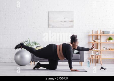 vista laterale della vivace afroamericana più dimensioni donna in abbigliamento sportivo che si esercita sul tappetino fitness Foto Stock
