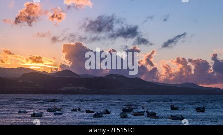 Paesaggio con piccole barche che galleggiano in mare al tramonto con il sole che tramonta dietro alcune montagne sullo sfondo Foto Stock