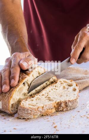 immagine verticale delle mani dell'uomo che rompono il pane organico nel primo piano con coltello circondato da briciole su tavola bianca con luce naturale Foto Stock