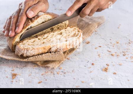 immagine orizzontale delle mani dell'uomo che rompono il pane organico nel primo piano con coltello circondato da briciole su tavola bianca con luce naturale Foto Stock