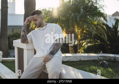 Uomo europeo sconvolto in una camicia bianca con braccia tatuate seduti all'aperto Foto Stock